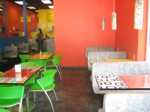 Seating area at Big Mouth Subs. Bright orange walls, bright green chairs, multicolored tables, and paint-spattered lighting fixtures.